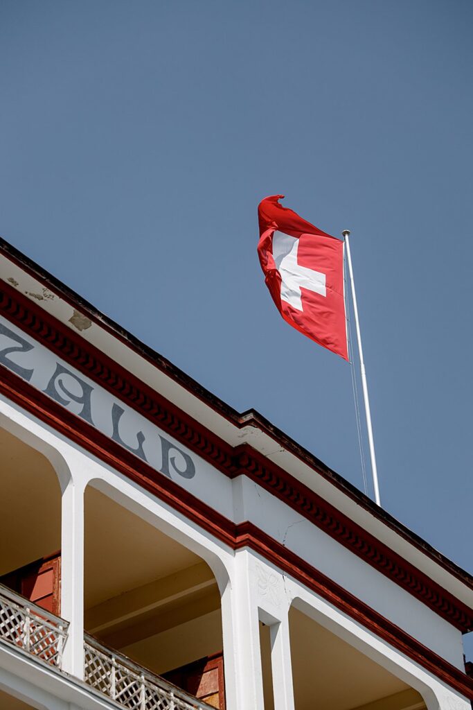 Hochzeit in den Schweizer Alpen 2 Die Schweizer Fahne weht an einem Fahnenmast auf einem weißen Gebäude mit roter Umrandung vor einem strahlend blauen Himmel - eine perfekte Szene für eine Luxus-Hochzeit im Ausland oder die Kulisse für einen unvergesslichen Film.