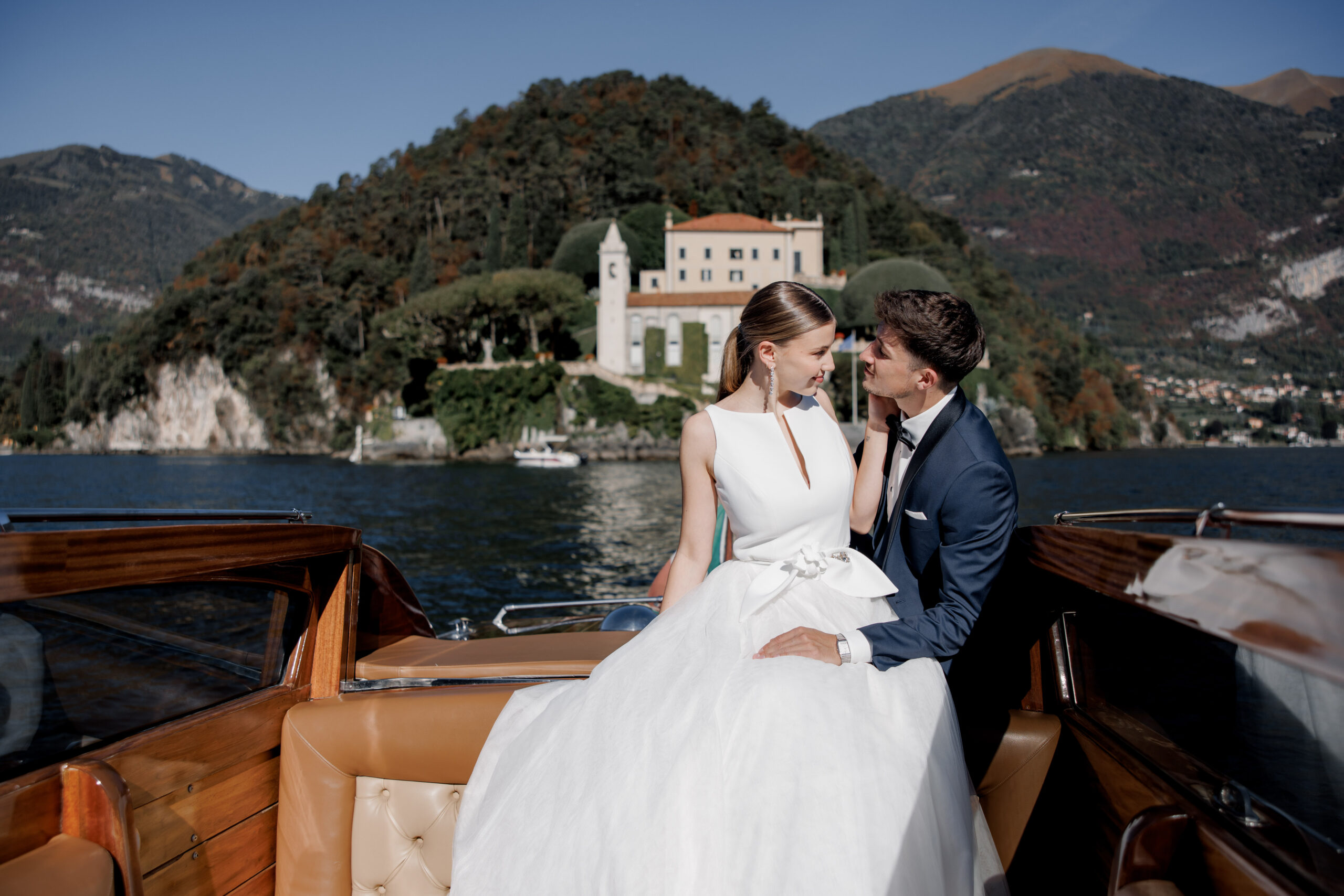 Eine Braut und ein Bräutigam lächeln sich auf einem Boot an, mit einer Villa und grünen Hügeln im Hintergrund, die die Essenz von Luxus für ihre unvergessliche Hochzeit im Ausland einfangen. Dieser Moment sieht aus wie eine Szene aus einem romantischen Film.