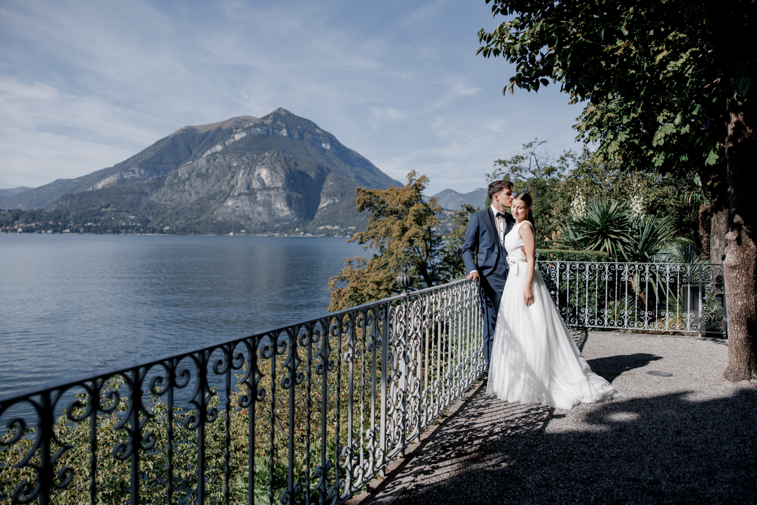 Die Braut und der Bräutigam posieren auf einer Seeterrasse mit den Bergen und dem blauen Himmel im Hintergrund und fangen so die Essenz von Luxus Fotografie bei einer Hochzeit im Ausland ein.