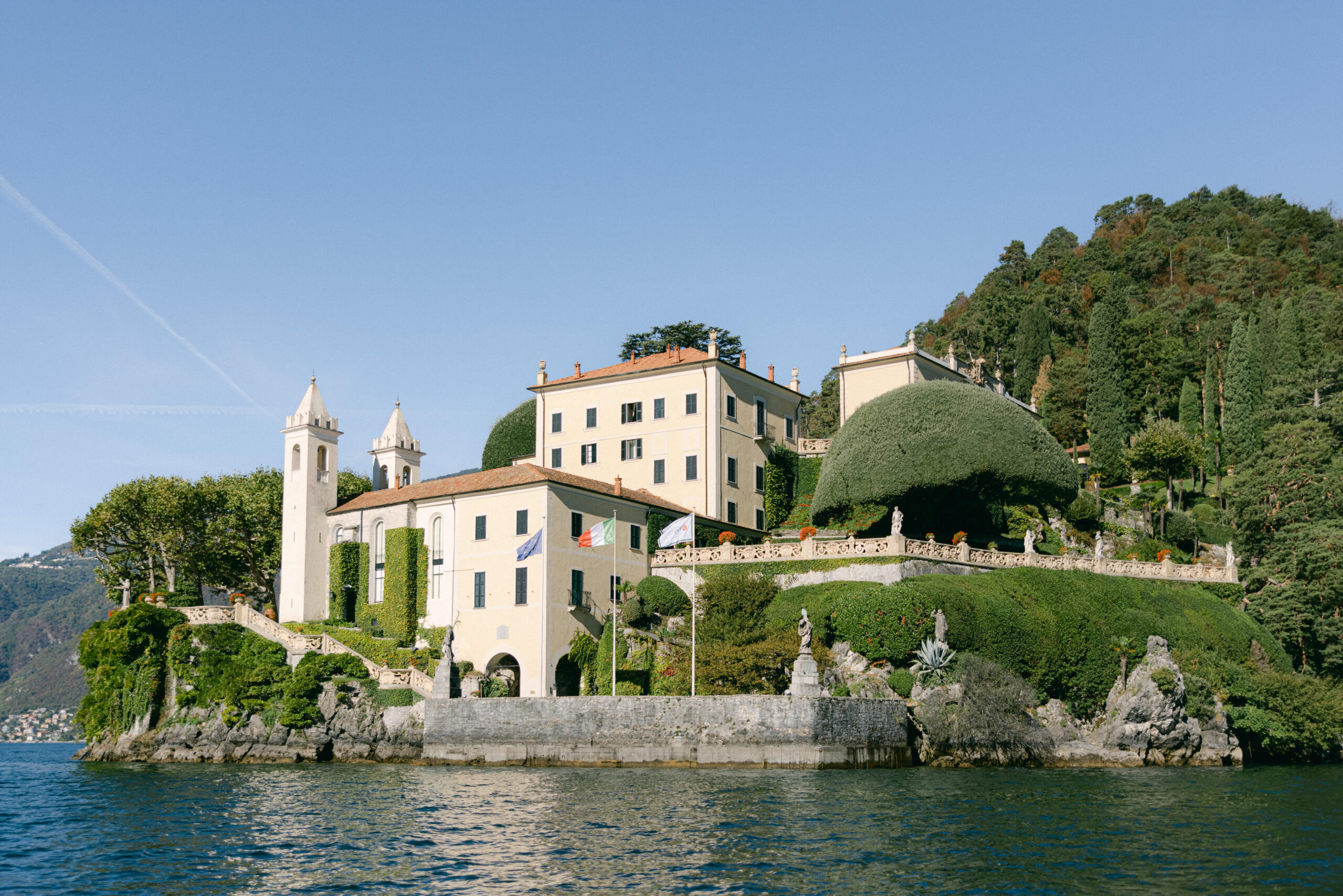 Elegante Villa mit gepflegtem Garten auf einem Hügel am See, perfekt für eine filmreife Hochzeit im Ausland, umgeben von Wasser und üppigem Grün unter einem klaren Himmel.