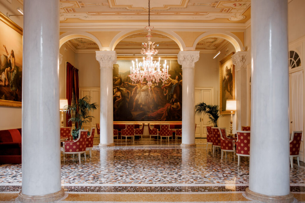 Die 10 schönsten Hochzeitslocations am Comer See 2 Elegante Hotellobby mit Marmorsäulen, Kronleuchtern, roten Stühlen und großen klassischen Gemälden an den Wänden - ein wahres Luxusziel, das direkt aus einem klassischen Film entsprungen scheint.