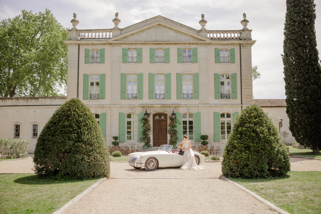 Braut und Bräutigam stehen in einem weißen Oldtimer vor einem prächtigen Schloss mit grünen Fensterläden und fangen die Eleganz einer Luxushochzeit im Ausland ein.