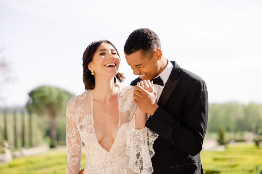 Ein fröhliches Brautpaar lacht an seinem Hochzeitstag im Freien, hält sich an den Händen und lächelt - ganz im Stil von Luxus Fotografie.