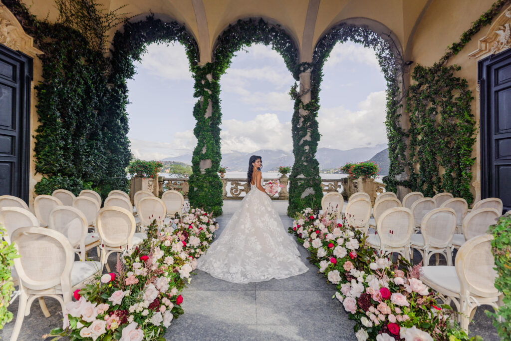 Die Braut in einem weißen Kleid steht in einem blumengeschmückten Hochzeitsort im Freien mit Bögen und einer malerischen Aussicht und fängt die Essenz von Luxus und eleganter Zielort-Romantik ein.
