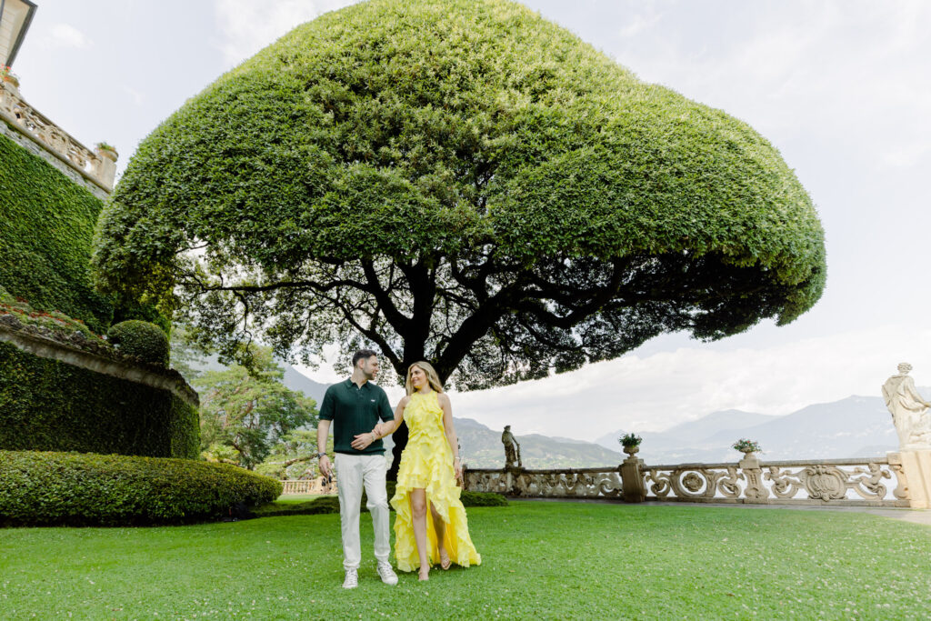 Ein Paar genießt einen luxuriösen Spaziergang auf saftig grünem Gras unter einem großen Baum in einem malerischen Garten, mit den Bergen im Hintergrund - das perfekte Ziel für eine Hochzeit im Ausland.