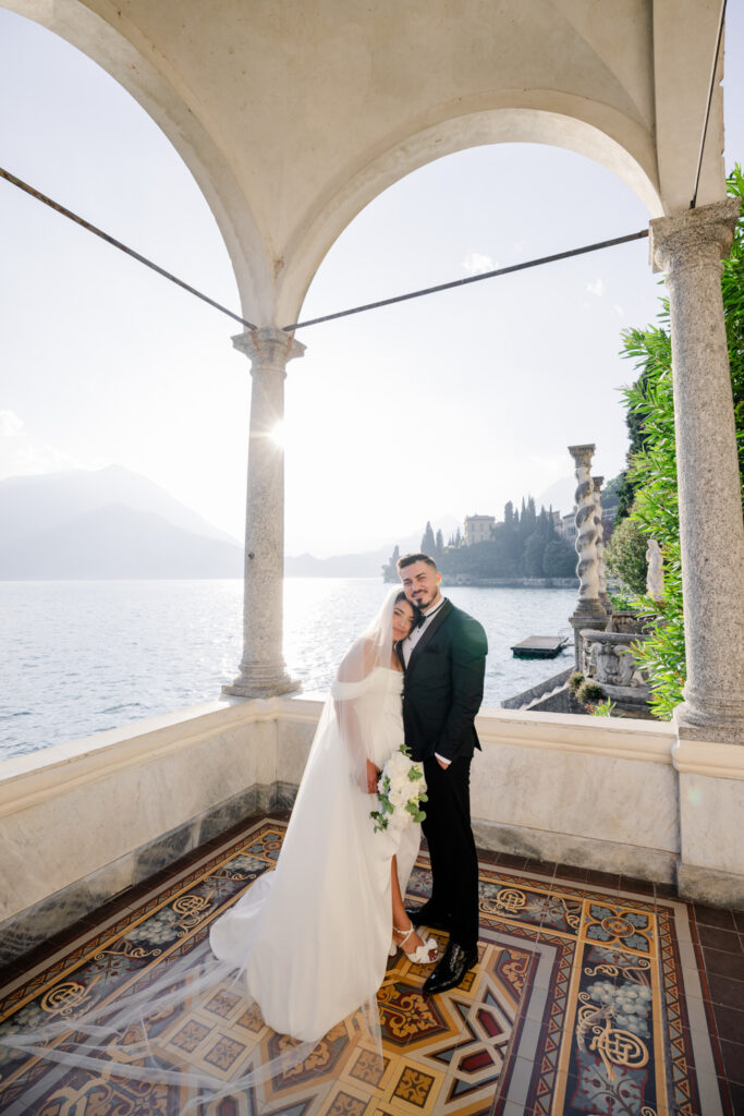 Die 10 schönsten Hochzeitslocations am Comer See 19 Eine Braut und ein Bräutigam lächeln sich unter einem eleganten Torbogen an einem luxuriösen Reiseziel am See zu, mit den Bergen im Hintergrund - eine Szene wie aus einem romantischen Film.