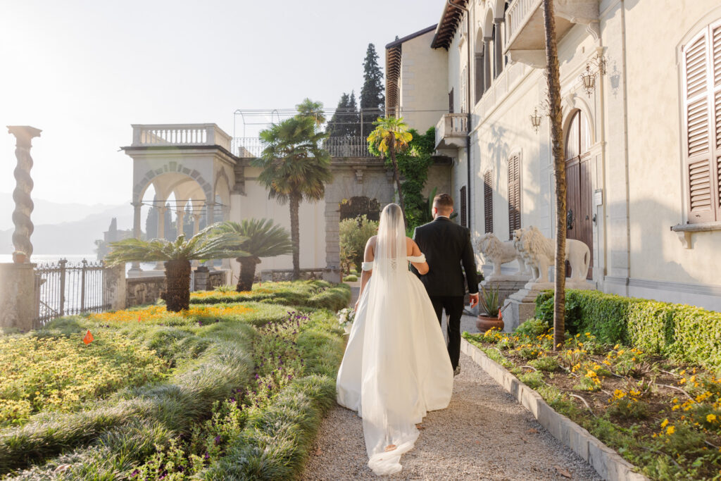 Die 10 schönsten Hochzeitslocations am Comer See 17 Braut und Bräutigam spazieren an einem sonnigen Tag einen Gartenweg neben einem historischen Gebäude entlang und fangen damit das Gefühl einer Luxus-Hochzeit wie aus einem Film ein.