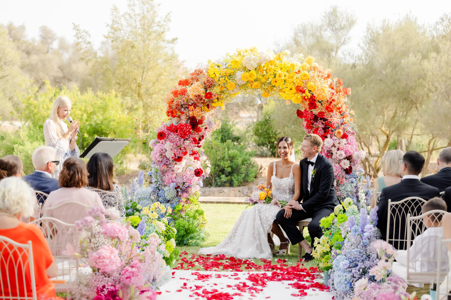 Wedding Photography Mallorca 17 Braut und Bräutigam sitzen unter einem farbenfrohen Blumenbogen bei einer luxuriösen Hochzeitszeremonie am Reiseziel, während die Gäste zuschauen, eingefangen in atemberaubender Fotografie.