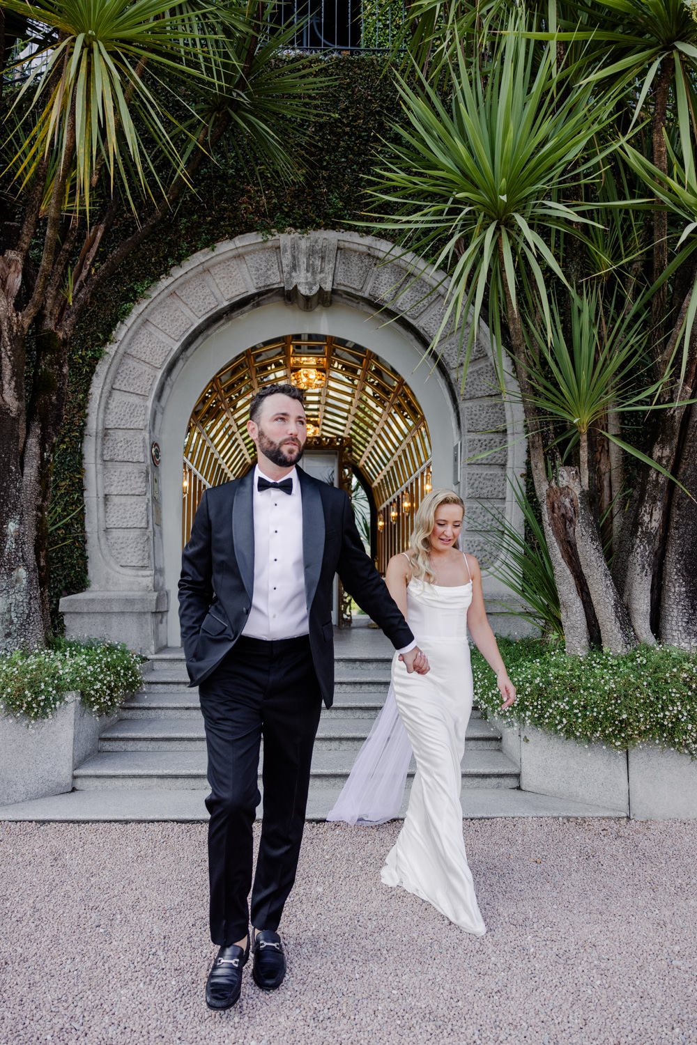 Ein Mann im Smoking und eine Frau im weißen Kleid schreiten Hand in Hand vor einem verzierten Torbogen, der den Luxus einer Hochzeit im Ausland einfängt wie eine Szene aus einem romantischen Film.