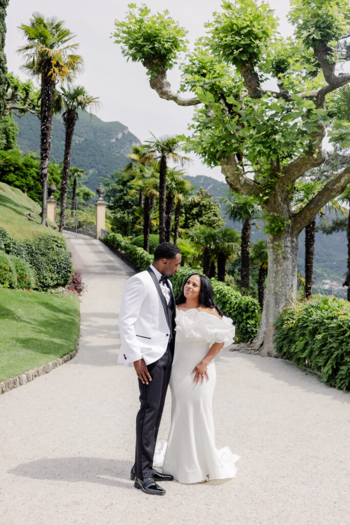 Ein formell gekleidetes Paar lächelt sich auf einem von Bäumen gesäumten Weg mit den Bergen im Hintergrund an und fängt die Romantik einer Hochzeit im Ausland durch zeitlose Fotografie ein.