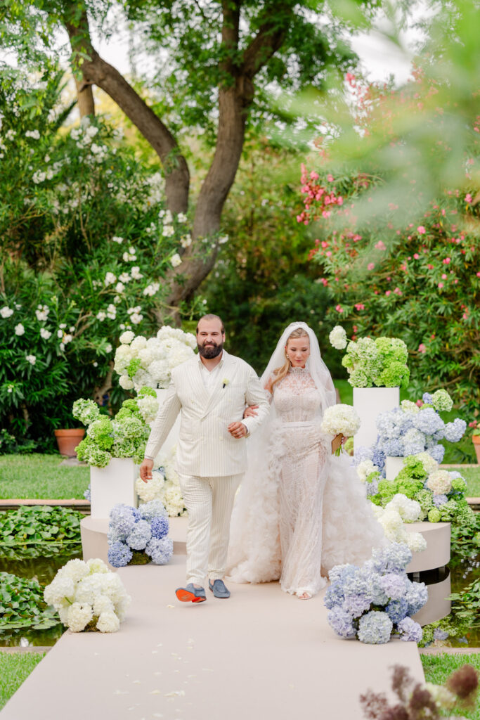 Braut und Bräutigam schreiten durch einen mit weißen und blauen Blumen gesäumten Gang im Freien, umgeben von üppigem Grün, und schaffen so eine filmreife Kulisse für ihre Hochzeit am Urlaubsort.