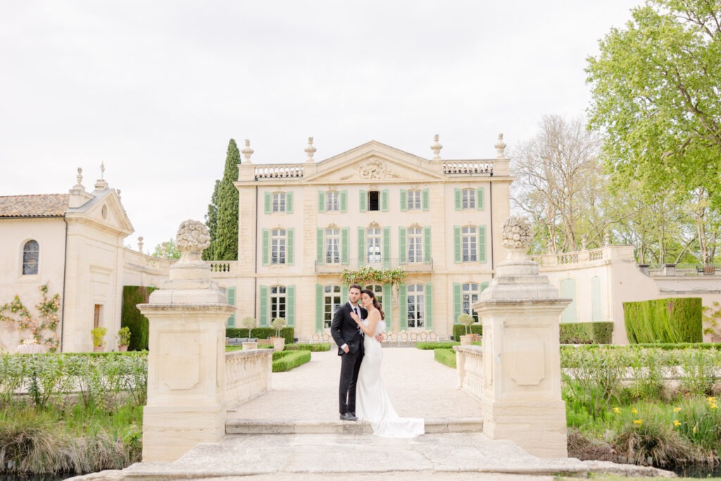 Bride and groom stand together in front of a large, elegant castle surrounded by greenery, capturing the magic of their destination wedding in timeless photography.