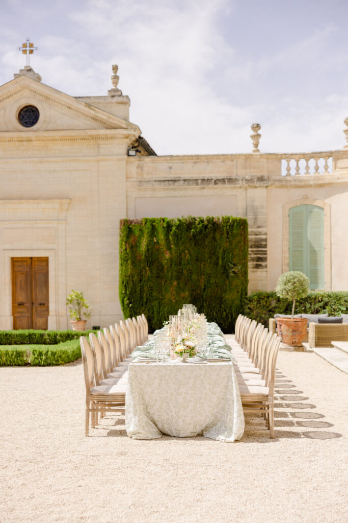 Long banquet table with elegant place settings in a sunny, historic courtyard, perfect for a luxury destination wedding, with a charming stone building as a backdrop.
