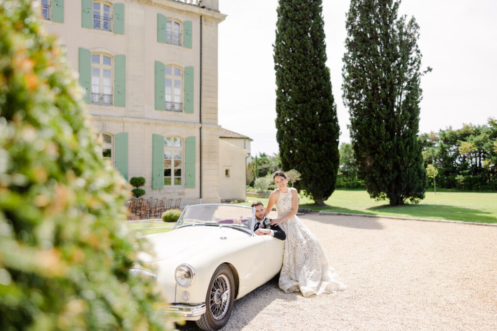Bride and groom pose with a classic white convertible in front of a large, elegant, green-covered building, capturing the essence of luxury destination photography.