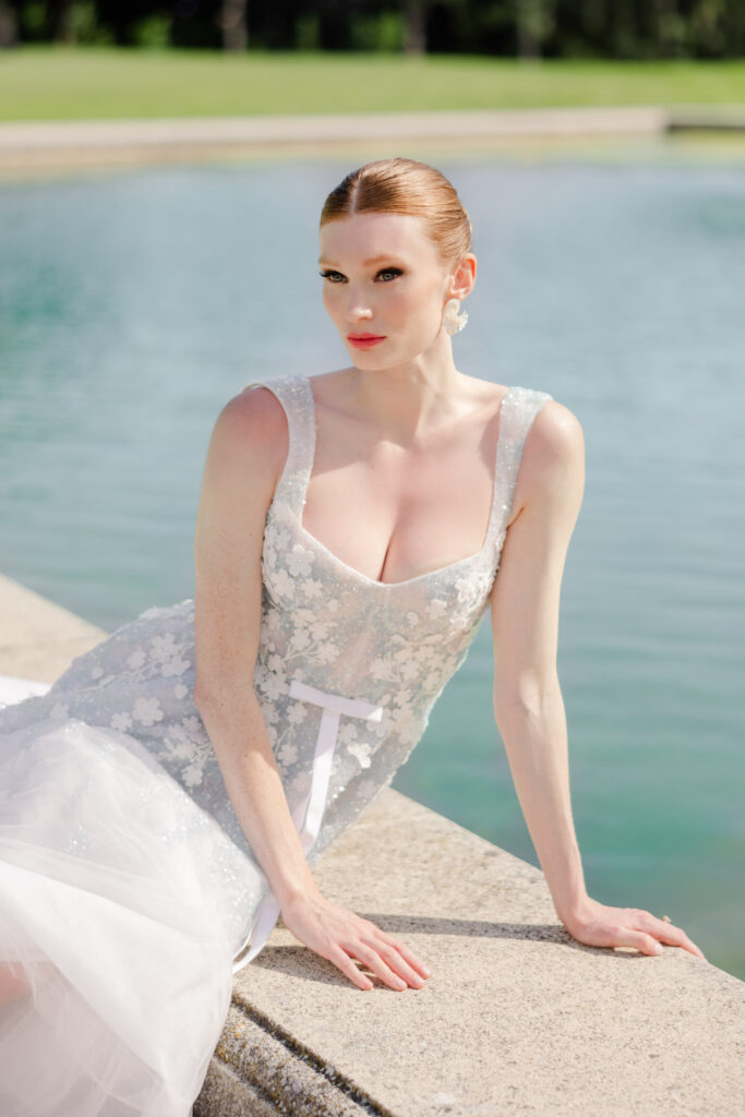 A woman in a white floral dress sits by a calm lake and gazes into the distance on a sunny day, reminiscent of the dreamy aesthetic of film photography.