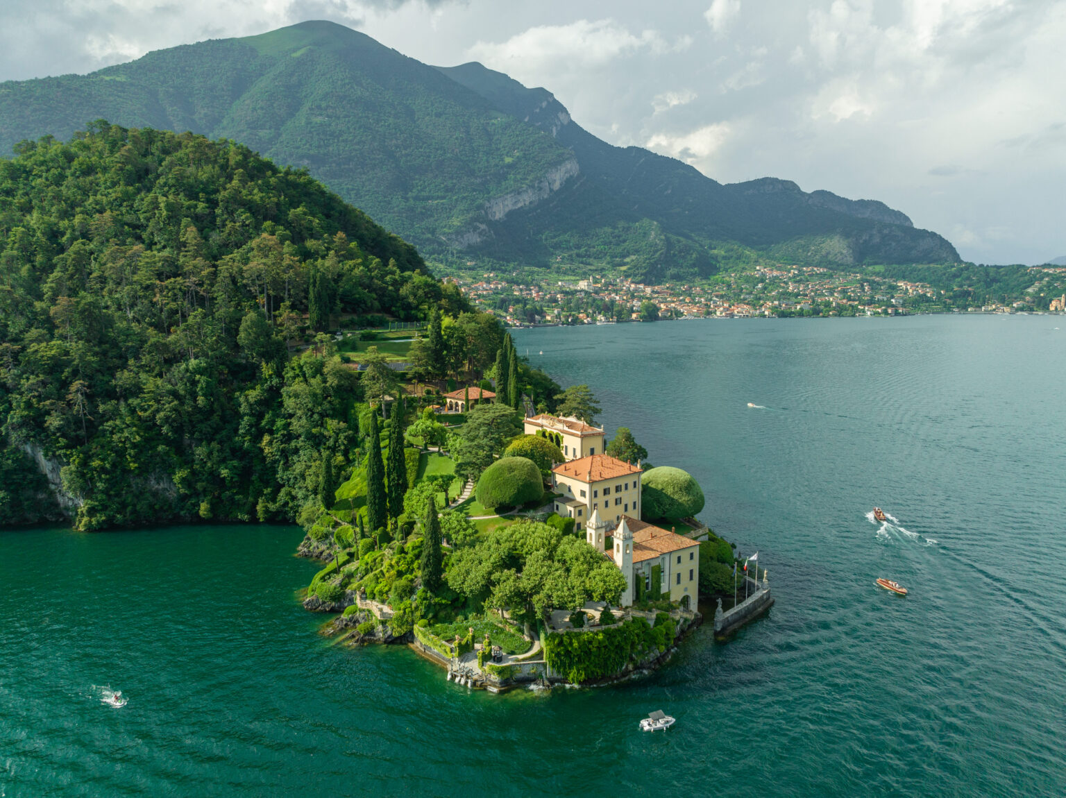 Drohnenaufnahme einer historischen Villa mit Parkanlage auf einer bewaldeten Halbinsel im Comer See vor Bergkulisse.