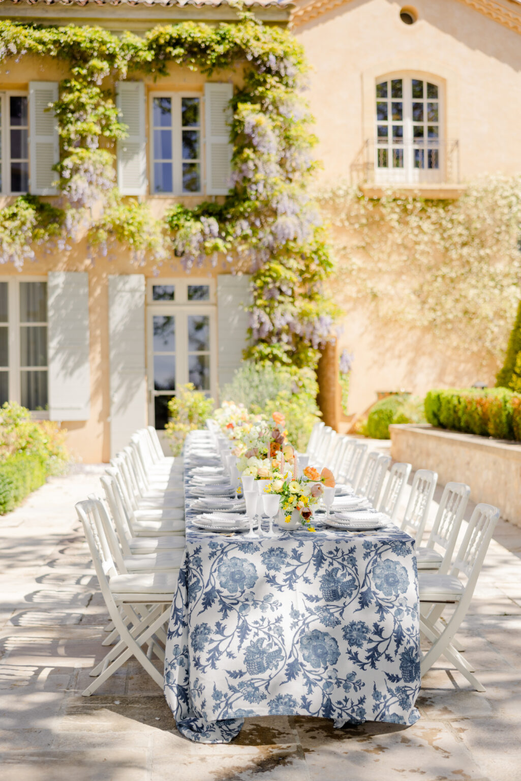 Wedding Photography South France 23 Long outdoor table with white chairs and a floral tablecloth, perfect for a destination wedding, set for a meal next to a villa covered in grapevines—ideal for unforgettable film and photo moments.
