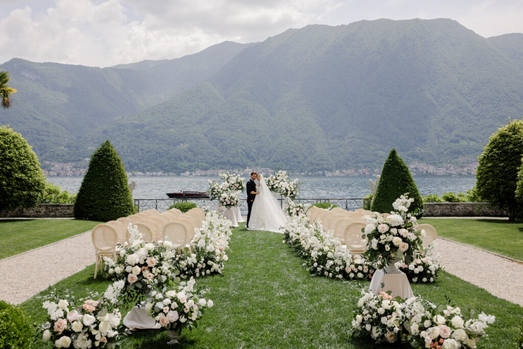 High-end luxury destination wedding photography at Lake Como, Italy, featuring an elegant outdoor ceremony by the lake with abundant white florals, a bride and groom, and dramatic mountain views, photographed by The Tailors Photography.