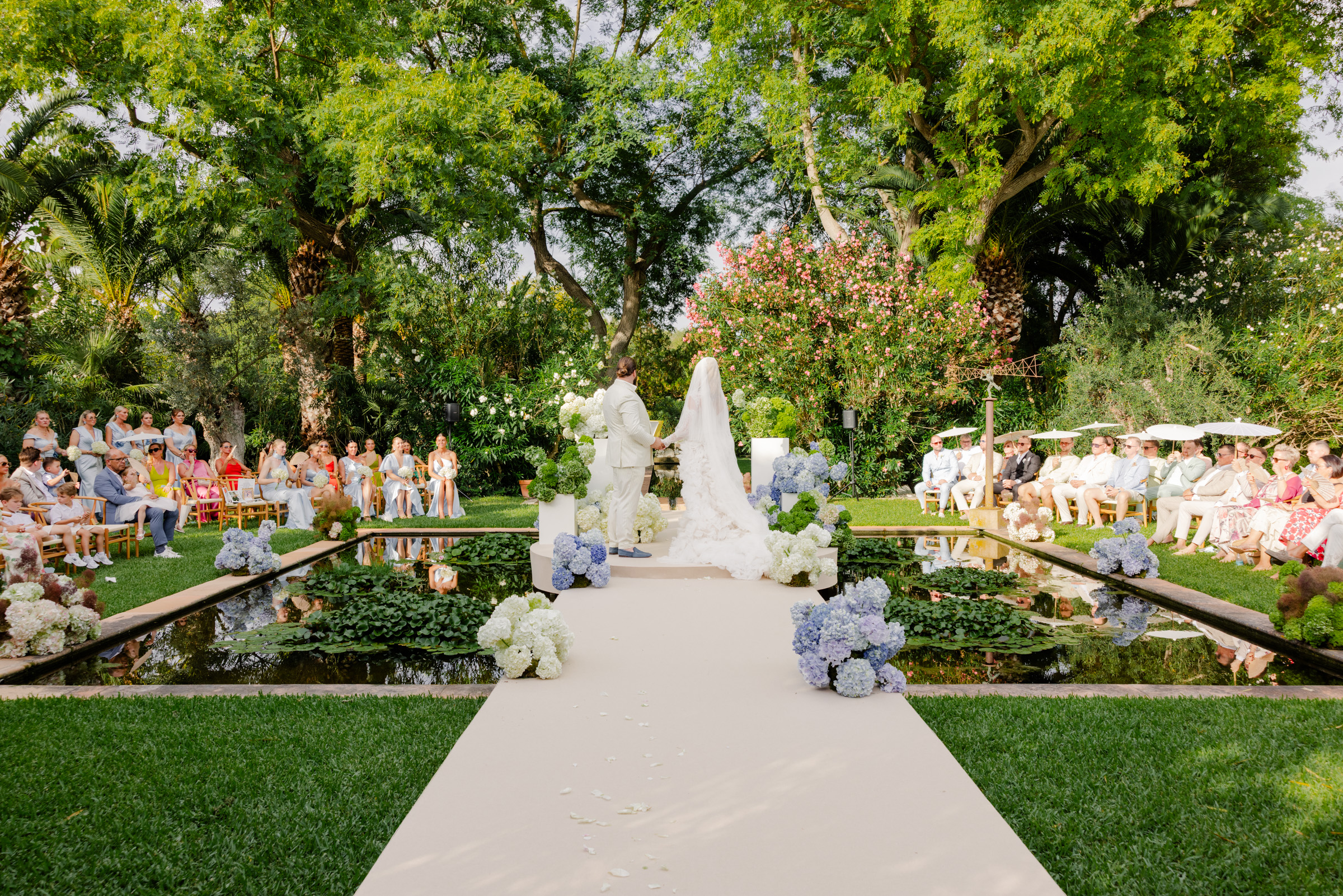 Eine Braut und ein Bräutigam stehen vor einem Hochzeitsaltar im Freien, umgeben von Gästen und üppigem Grün.
