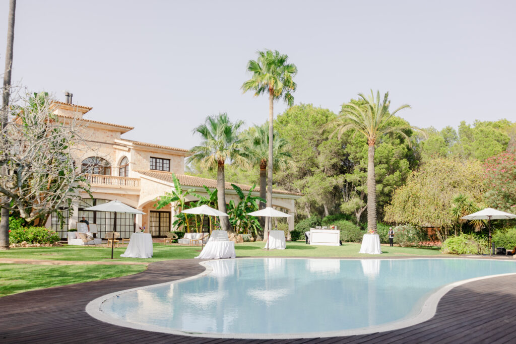 Elegante Villa mit Palmen, Pool und Tischen mit weißen Sonnenschirmen, die auf einem grünen Rasen für eine Veranstaltung im Freien aufgestellt sind.