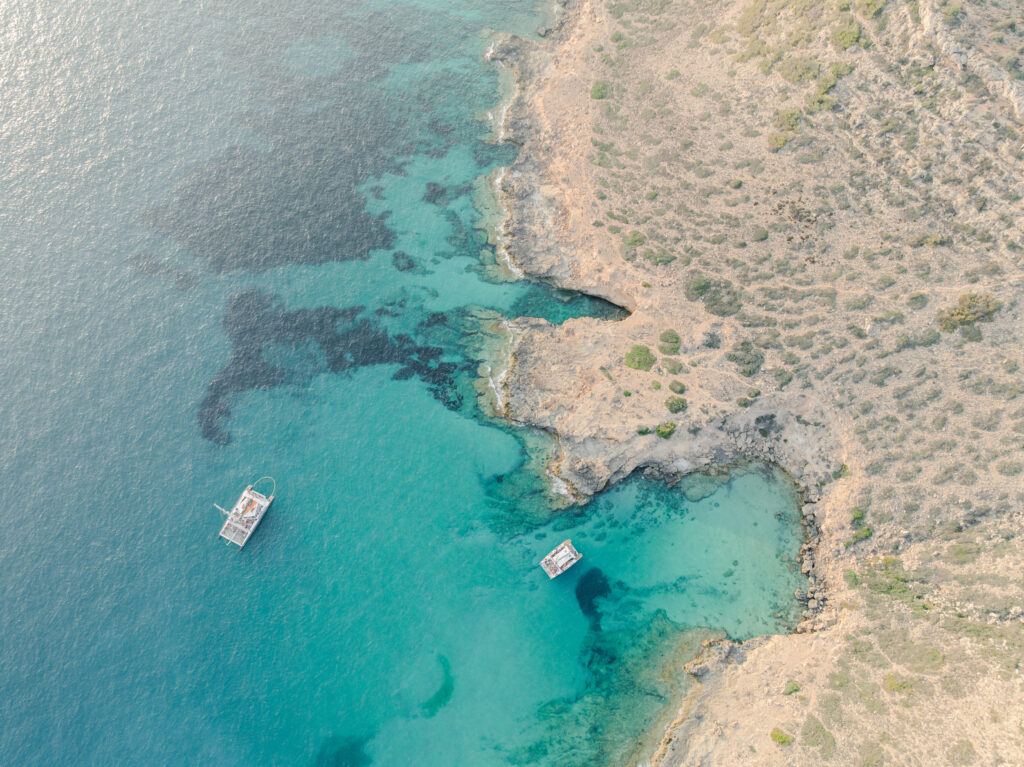 Luftaufnahme von zwei Booten in klarem türkisfarbenem Wasser nahe einer felsigen, sonnenbeschienenen Küste mit spärlicher Vegetation.