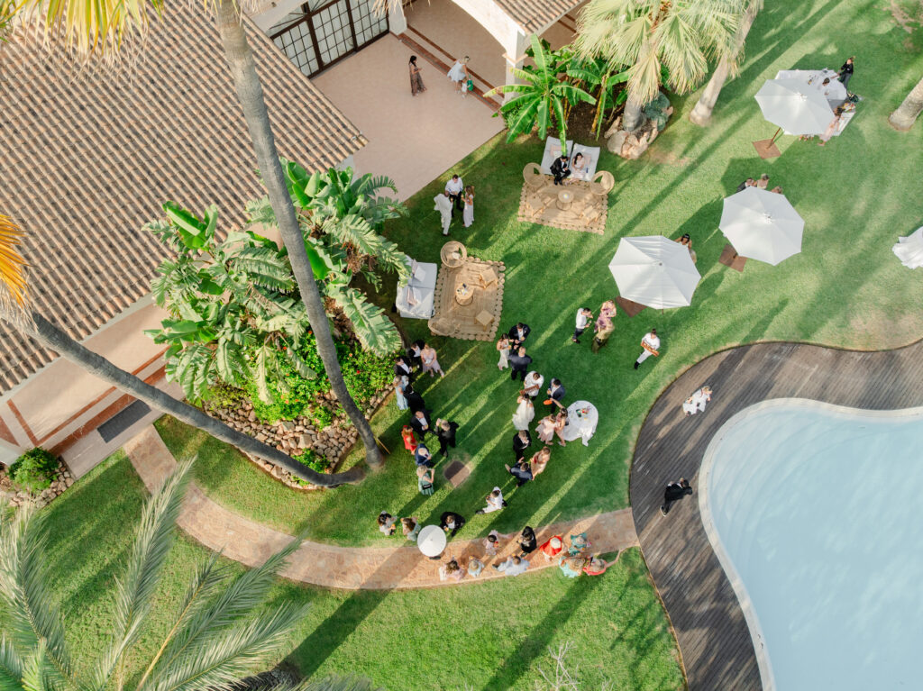 Luftaufnahme von Menschen, die bei einer Veranstaltung im Freien mit Sonnenschirmen und Palmen in der Nähe eines Pools und eines Gartens versammelt sind.