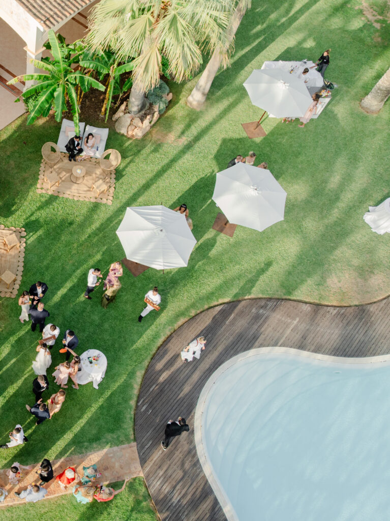 Luftaufnahme von Menschen, die an einem sonnigen Tag an einem Pool mit weißen Sonnenschirmen und Palmen sitzen.