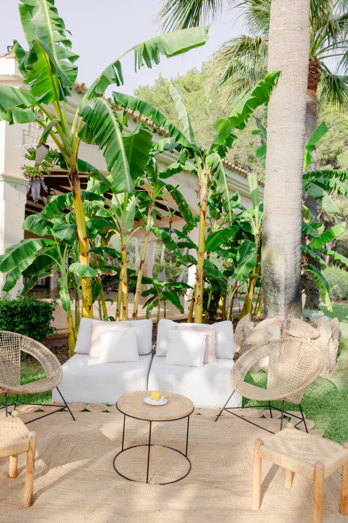 Sitzbereich im Freien mit weißen Kissen, Korbstühlen und einem runden Tisch, umgeben von Palmen und Bananenbäumen.