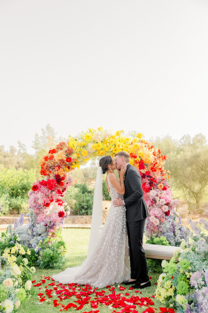 Braut und Bräutigam küssen sich unter einem leuchtenden Regenbogen im Freien, umgeben von bunten Blumen.