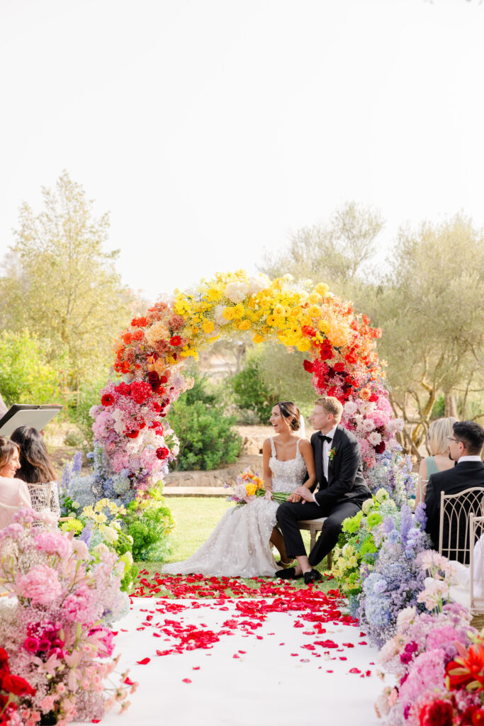 Braut und Bräutigam sitzen bei einer Hochzeitszeremonie im Freien unter einem bunten Blumenbogen, umgeben von Blumenarrangements.