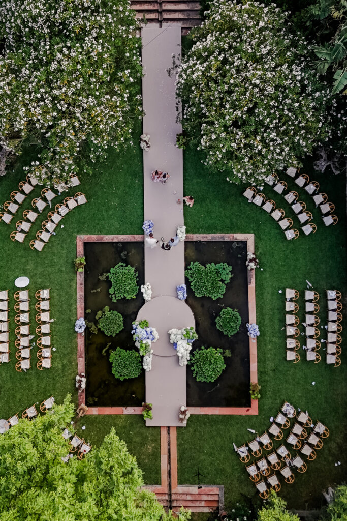 Luftaufnahme einer Hochzeit im Freien mit Stühlen, Bäumen und einem Gartenweg, der eine Kreuzform bildet.