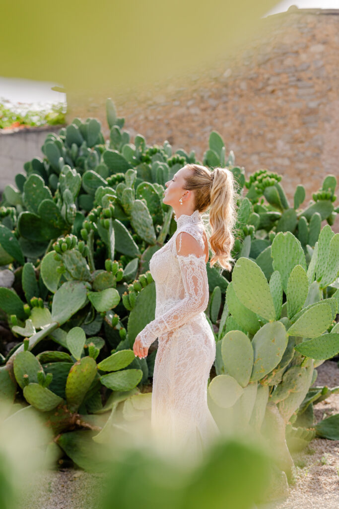 Eine Frau in einem Spitzenkleid steht inmitten grüner Kakteen und blickt zur Seite, während das Sonnenlicht ihr blondes Haar beleuchtet.