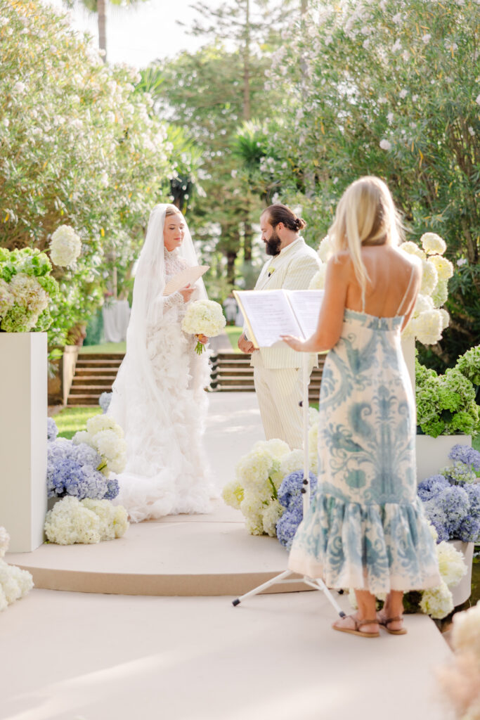Braut und Bräutigam stehen gemeinsam im Freien, umgeben von Blumen, während ein Zeremonienmeister aus einer Broschüre liest.