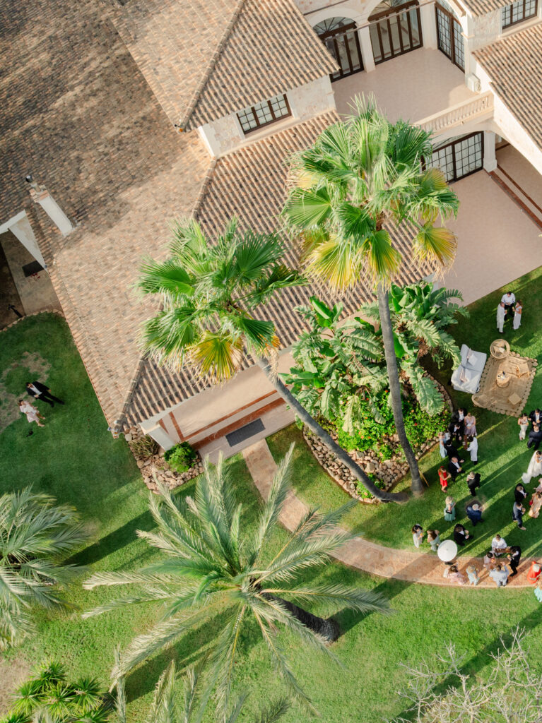 Luftaufnahme von Menschen, die sich in einem Garten in der Nähe eines großen Hauses mit Palmen und grünen Rasenflächen versammelt haben.