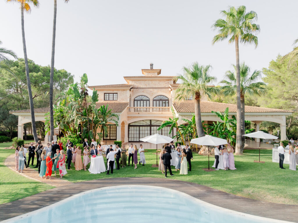 Eine Gruppe von formell gekleideten Personen versammelt sich an einem sonnigen Tag an einem Pool und einer großen Villa, die von Palmen umgeben ist.