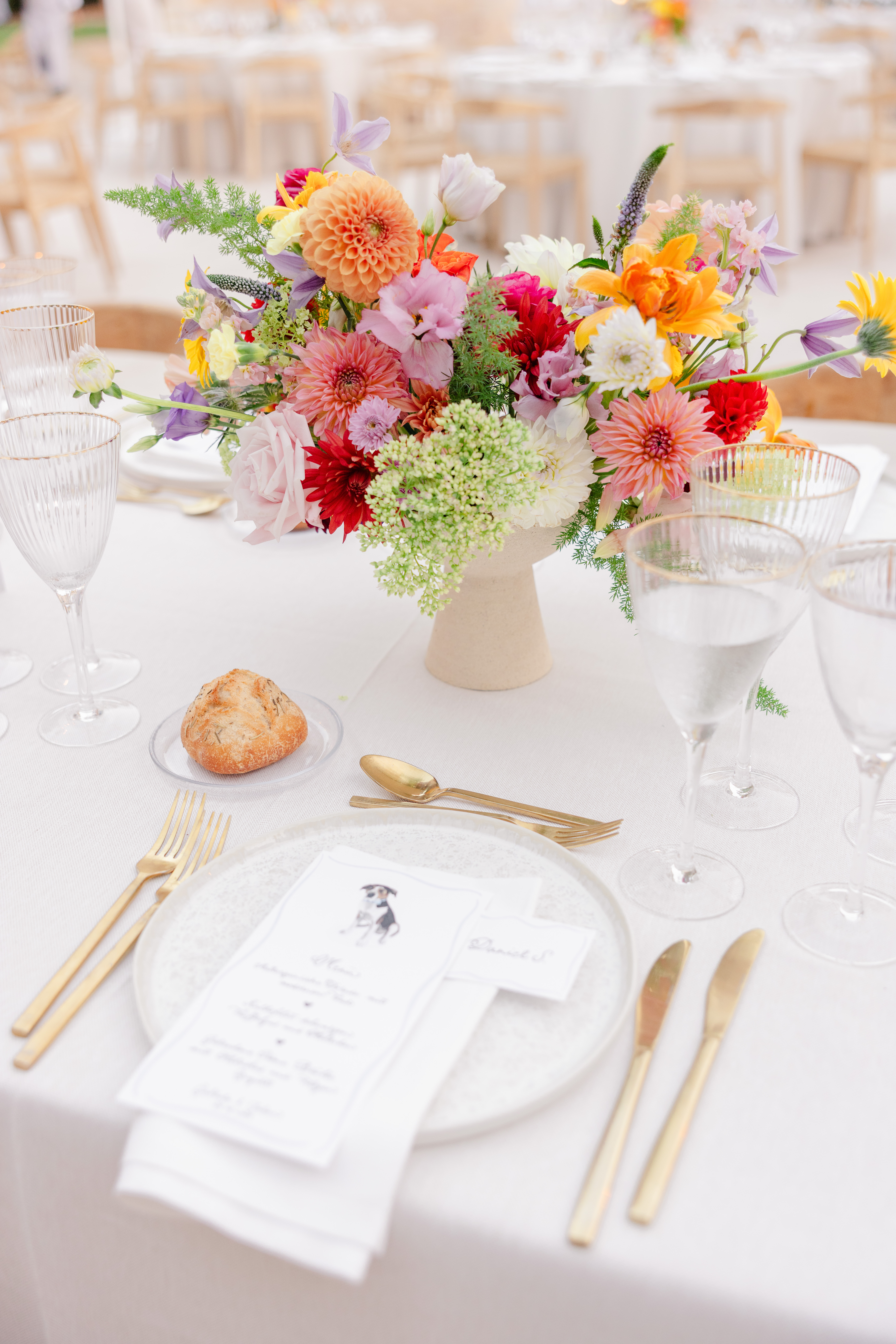 Wedding Photography Mallorca 19 Elegant gedeckter Tisch mit goldenen Utensilien, einer Speisekarte, einem Brötchen und einem farbenfrohen Blumenschmuck in der Mitte.