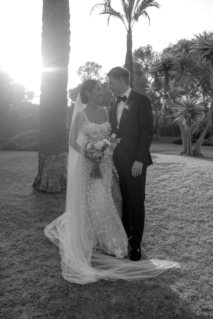 Eine Braut und ein Bräutigam stehen zusammen im Freien und lächeln sich an, während das Sonnenlicht durch die Bäume scheint.