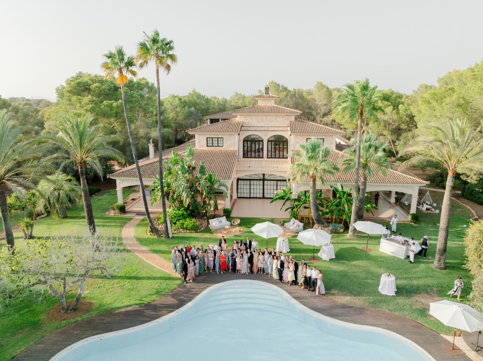 Wedding Photography Mallorca 22 Eine große Gruppe versammelt sich an einem Pool vor einer Villa, die von Palmen und Grünflächen umgeben ist.