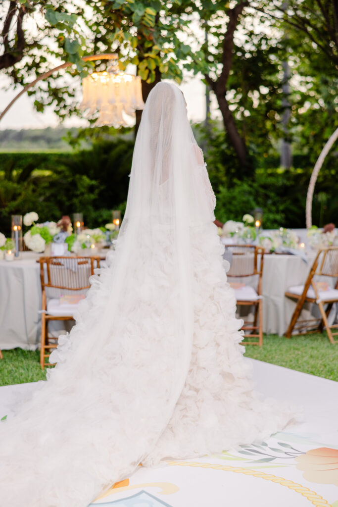 Eine Braut in einem langen, weißen Rüschenkleid und Schleier steht im Freien neben geschmückten Tischen und Holzstühlen.