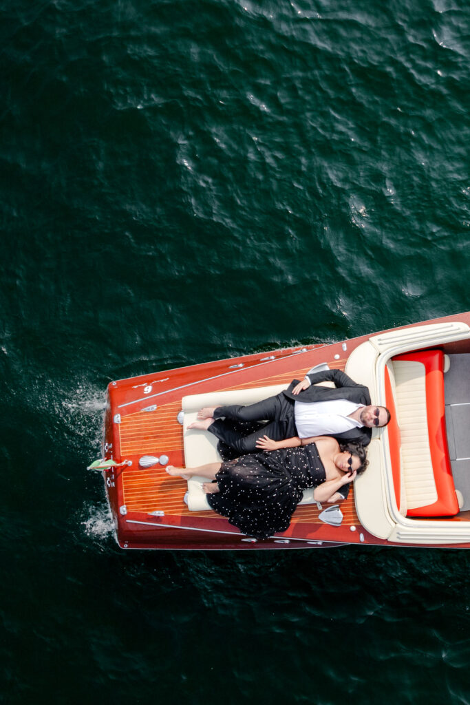 Ein formell gekleidetes Paar entspannt sich auf einem Holzboot, das auf tiefgrünem Wasser schwimmt, von oben gesehen.
