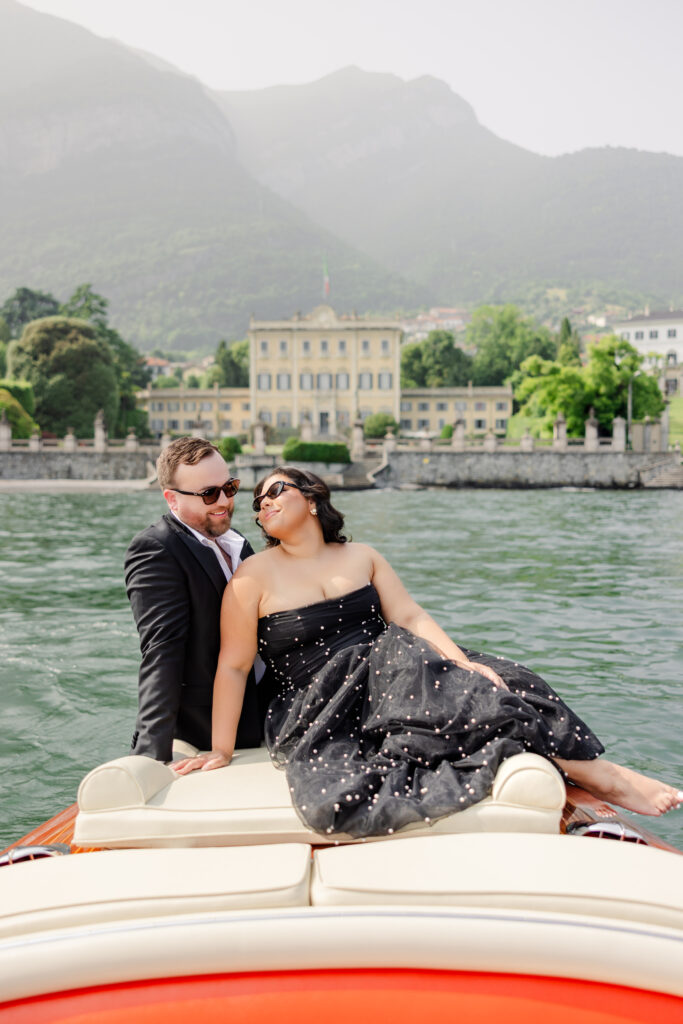 Ein Paar in formeller Kleidung entspannt sich auf einem Boot mit einer großen Villa und Bergen im Hintergrund.