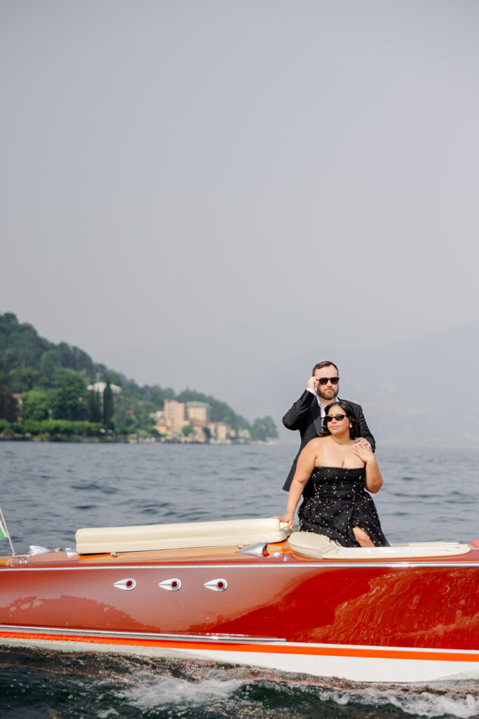 Ein stilvolles Paar in formeller Kleidung fährt auf einem roten Schnellboot auf einem See mit Hügeln und Gebäuden im Hintergrund.