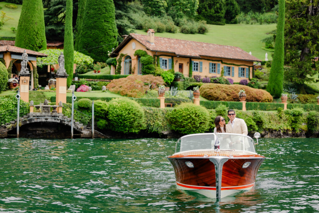 A couple rows a small wooden boat on a lake in front of a villa surrounded by lush greenery.