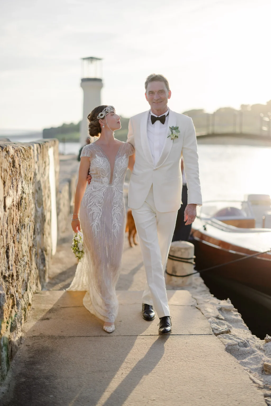A couple walks arm in arm along a stone path next to a lighthouse and a boat.