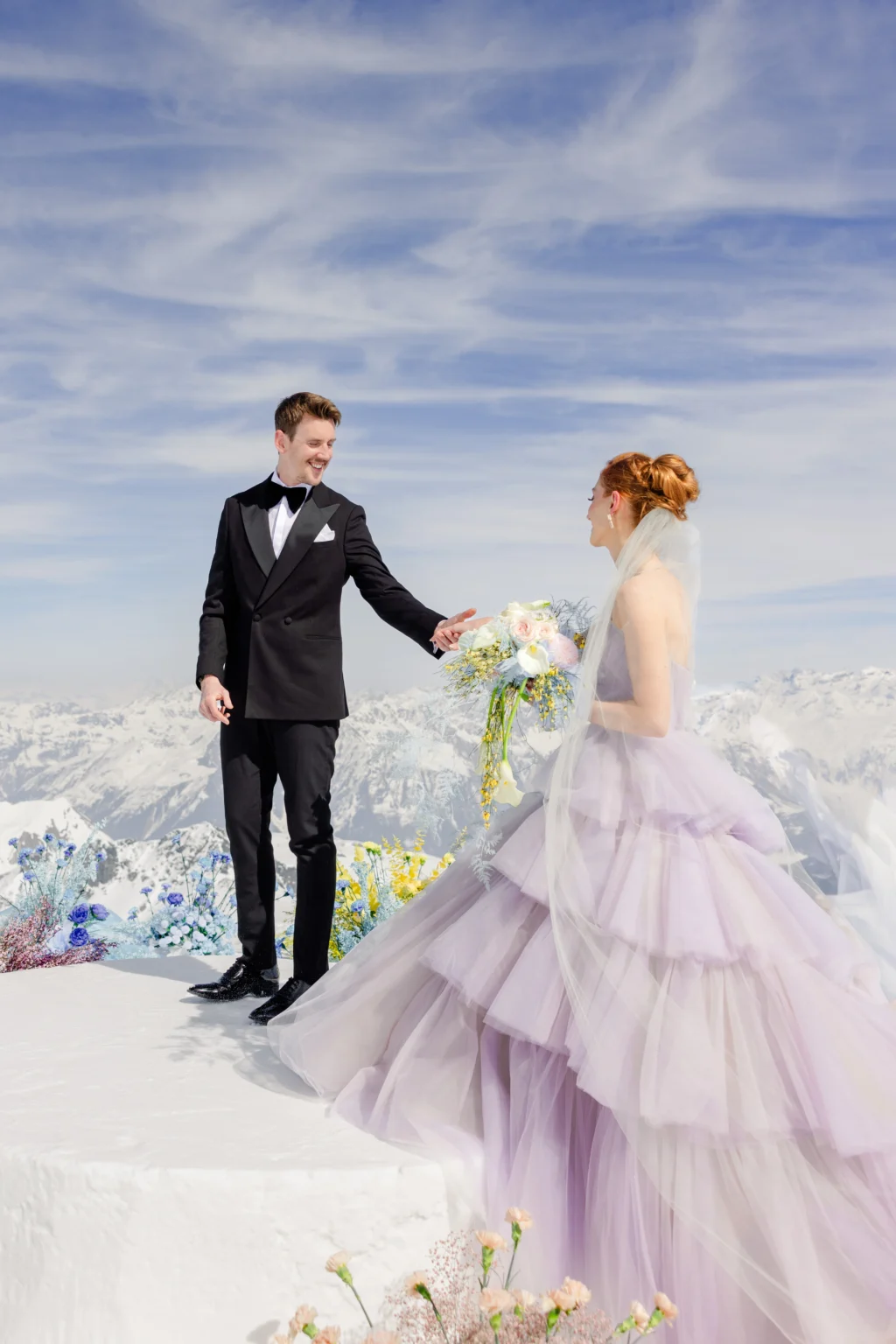 A bride in a lavender-colored, pleated wedding dress and a groom in a tuxedo stand on a snow-covered mountain peak.