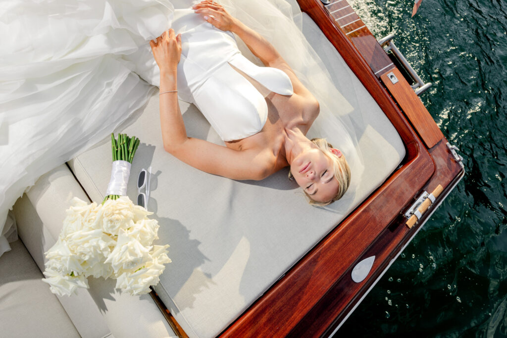 Hochzeitslocation Relais Villa Vittoria am Comer See – Intime Hochzeit in einer historischen Villa 22 Eine Braut in einem weißen Kleid liegt auf dem Deck eines Holzbootes neben einem Strauß weißer Rosen.