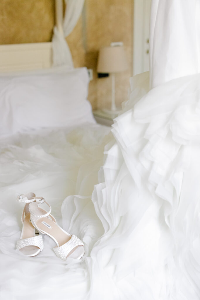 Hochzeitslocation Relais Villa Vittoria am Comer See – Intime Hochzeit in einer historischen Villa 8 Weiße Brautpumps, die auf einem Brautkleid auf einem Bett im Relais Villa Vittoria liegen.